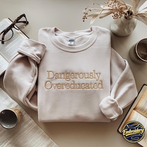 May include: A light beige crewneck sweatshirt featuring the embroidered text "Dangerously Overeducated" in a dark tan thread across the chest. The garment has long sleeves with ribbed cuffs and a ribbed neckline, presented in a neutral tone.