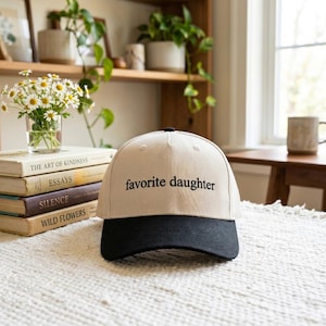 May include: A beige baseball cap with a black brim. The text "favorite daughter" is printed in black on the front of the cap. The cap is sitting on a white textured surface.