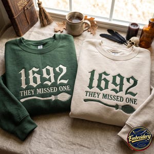 Peut inclure: Deux sweat-shirts à col rond, un vert foncé et un crème, arborant chacun une broderie avec le texte 1692 THEY MISSED ONE au-dessus d'un motif de balai. Le sweat-shirt vert présente une broderie crème, tandis que le modèle crème a une broderie vert foncé.
