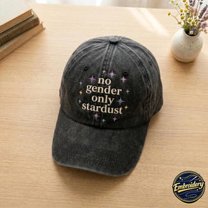 May include: A charcoal grey baseball cap with a distressed finish. The front features embroidered text that reads "no gender only stardust" in a light cream colour, surrounded by small, multi-coloured embroidered star icons in purple, blue, and yellow.