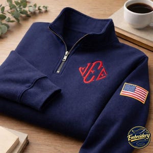 May include: A navy blue quarter-zip pullover sweatshirt featuring a red embroidered monogram on the chest and a rectangular American flag patch on the sleeve. The garment has a high collar with a silver-toned metal zipper closure.