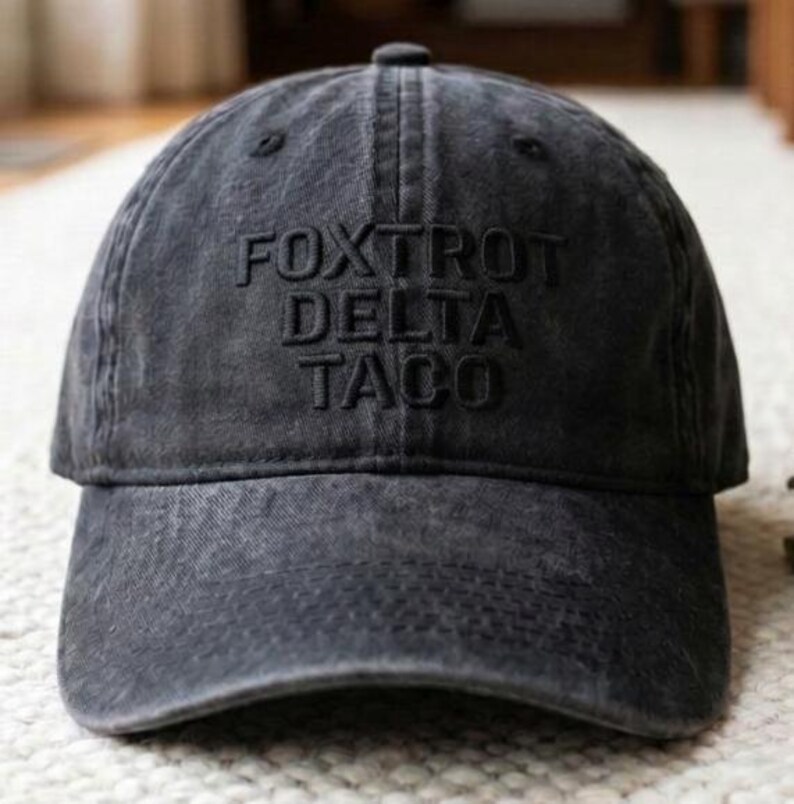 May include: A dark gray baseball cap with the words "FOXTROT DELTA TACO" embroidered on the front. The cap has a curved brim and a button on top. The fabric appears to be slightly faded, giving it a vintage look.