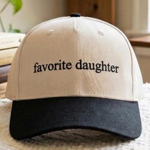 May include: A beige baseball cap with a black brim. The phrase "favorite daughter" is embroidered in black on the front of the cap. The cap has a curved brim and a button on top.
