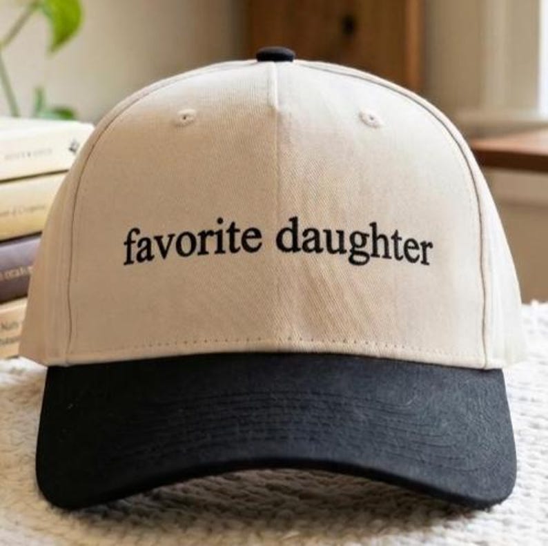 May include: A beige baseball cap with a black brim. The phrase "favorite daughter" is embroidered in black on the front of the cap. The cap has a curved brim and a button on top.