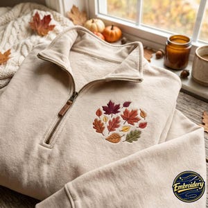 May include: A beige quarter-zip sweatshirt featuring an embroidered cluster of autumn leaves in shades of red, orange, yellow, and green on the chest. The garment has a metal zip pull and ribbed cuffs, showcasing a detailed autumn-themed design.