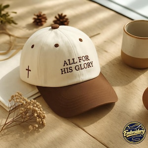 Puede incluir: Una gorra de béisbol color crema con visera marrón y un botón marrón en la parte superior. El frente presenta el texto bordado ALL FOR HIS GLORY en marrón, y una pequeña cruz marrón está bordada en el lateral. La gorra tiene ojales marrones.