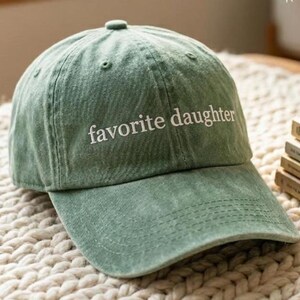 May include: A sage green baseball cap with the phrase "favorite daughter" embroidered in white. The cap has a curved brim and a classic six-panel design. The hat is made of a soft, textured fabric.