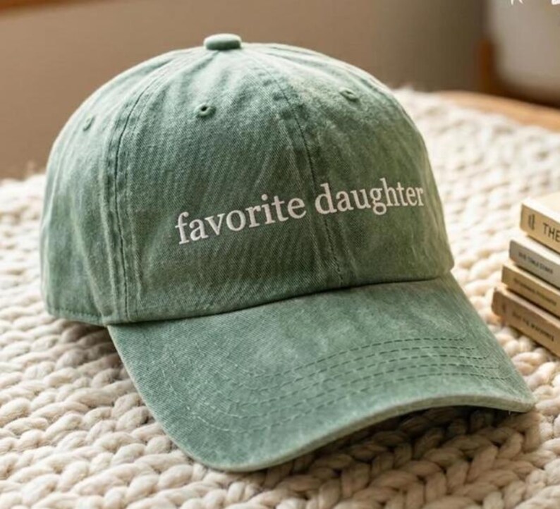 May include: A sage green baseball cap with the phrase "favorite daughter" embroidered in white. The cap has a curved brim and a classic six-panel design. The hat is made of a soft, textured fabric.