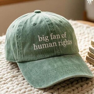 K&ouml;nnte beinhalten: Eine hellgr&uuml;ne Baseballkappe mit dem wei&szlig;en Schriftzug "big fan of human rights". Die Kappe hat einen gebogenen Schirm und einen Stoffriemenverschluss. Der Hut ist aus weichem, strukturiertem Material.