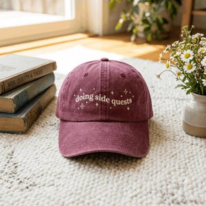 May include: A maroon baseball cap featuring the white embroidered text "doing side quests" centered on the front panel, surrounded by small white star-shaped icons. The cap has a structured crown, a curved brim, and visible stitching details.