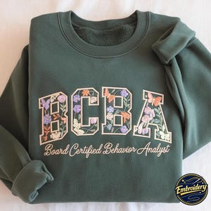 May include: A dark green crewneck sweatshirt featuring the embroidered letters BCBA in a collegiate font. The letters are filled with colourful floral and butterfly designs. Below the letters, the text Board Certified Behavior Analyst is embroidered.