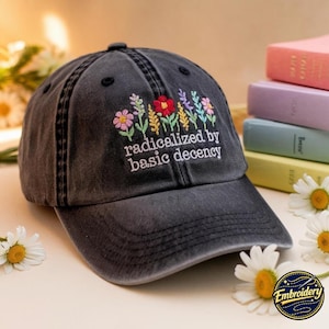May include: A charcoal grey baseball cap featuring embroidered colourful wildflowers and the white text "radicalized by basic decency" on the front panel. The cap has a curved brim and a distressed, washed-out fabric finish with visible stitching.