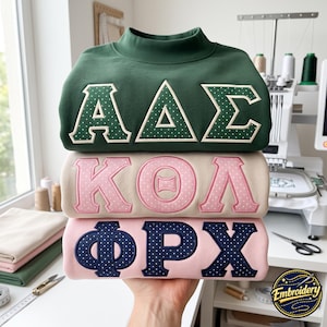 May include: A stack of three mock neck sweatshirts featuring Greek letters. The top is dark green with white polka-dot letters, the middle is beige with pink polka-dot letters, and the bottom is light pink with navy blue polka-dot letters.
