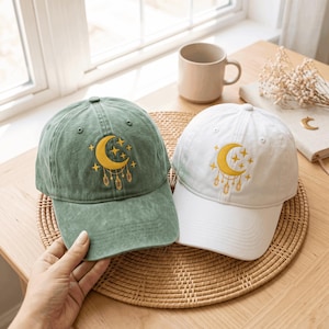May include: Two baseball caps, one in sage green and one in white, each featuring an embroidered yellow crescent moon, stars, and hanging crystal charms on the front panel. Both caps have a structured crown, a curved brim, and eyelet vents.