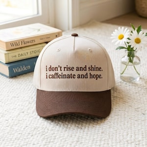 May include: A beige and brown baseball cap with the text "i don't rise and shine. i caffeinate and hope." The cap is sitting in front of a stack of books and a vase of daisies.