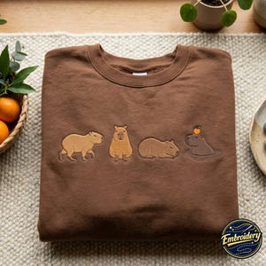 May include: A brown crewneck sweatshirt featuring an embroidered design of four capybaras in different poses. The first is walking, the second is sitting, the third is lying down, and the fourth is swimming in water with a small orange on its head.
