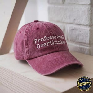 May include: A maroon baseball cap with a distressed, washed-out finish. The front of the cap features white embroidered text that reads Professional Overthinker in a clean, sans-serif font. The cap has a curved brim and a structured crown.
