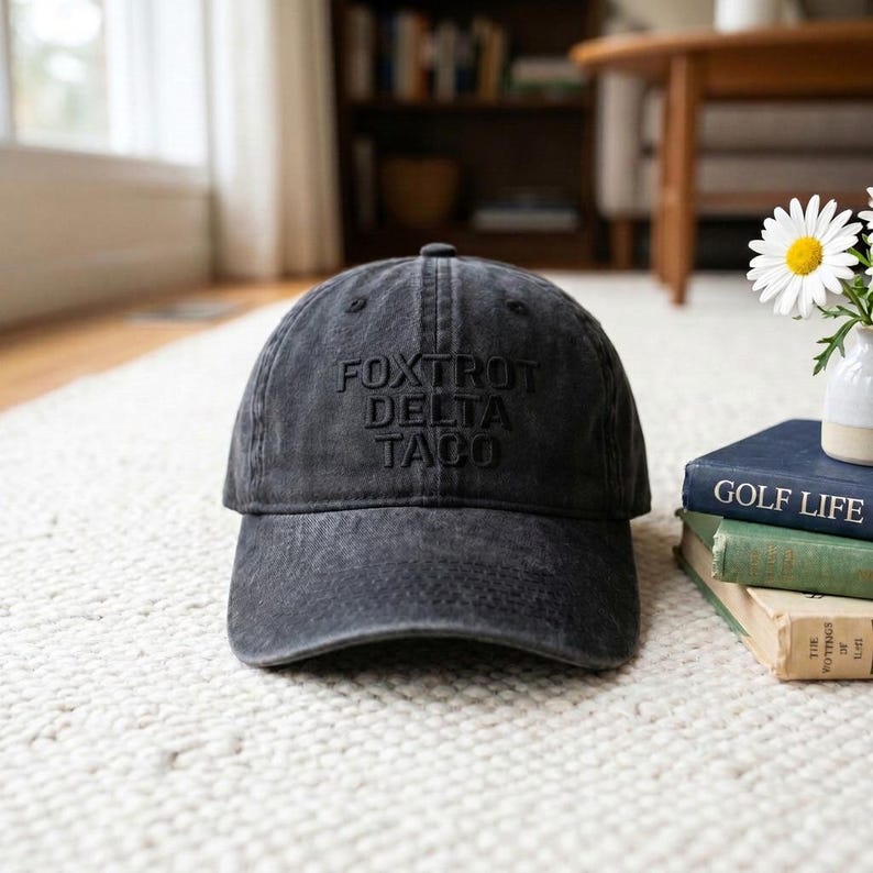 May include: A dark gray baseball cap with the words "FOXTROT DELTA TACO" embroidered on the front. The hat is sitting on a light-colored rug. The hat has a curved brim and a fabric strap closure.