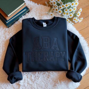 May include: A black sweatshirt with the words "ABA THERAPY" embroidered in a collegiate style. The sweatshirt is displayed on a white, fluffy rug with books and flowers in the background.