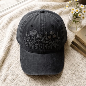 May include: A charcoal grey baseball cap featuring detailed tonal embroidery of various wildflowers across the front panels. The cap has a structured crown, a curved brim with stitched detailing, and a button at the top centre of the fabric panels.