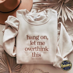 May include: A cream-coloured crewneck sweatshirt featuring the embroidered text "hang on, let me overthink this" in a dark brown thread. The garment has long sleeves with ribbed cuffs and a ribbed neckline, showcasing a clean and simple design.