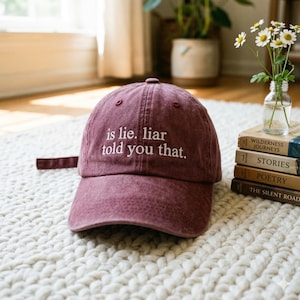 May include: A maroon baseball cap with the embroidered text "is lie. liar told you that." The cap has a curved brim and an adjustable strap. The hat is sitting on a white, textured surface.