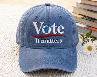 May include: A blue baseball cap with a distressed finish featuring white embroidered text that reads Vote with a blue checkmark replacing the letter o and a red curved line underneath followed by the white embroidered text It matters.