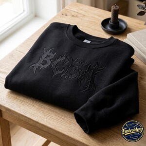 Op de afbeelding: Een zwart sweatshirt met ronde hals en ton-sur-ton borduursel op de borst. Het borduursel toont de letters BCBA in een gestileerd, scherp, vlamachtig lettertype. Het kledingstuk heeft lange mouwen met geribbelde boorden en een geribbelde hals.