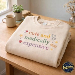 Może przedstawiać: Beżowa bluza z okrągłym dekoltem z kolorowym haftowanym napisem "cute and medically expensive". Wzór zawiera małe wyszywane ikony tabletki, stetoskopu, serca, plastra oraz kilku czteroramiennych gwiazdek.