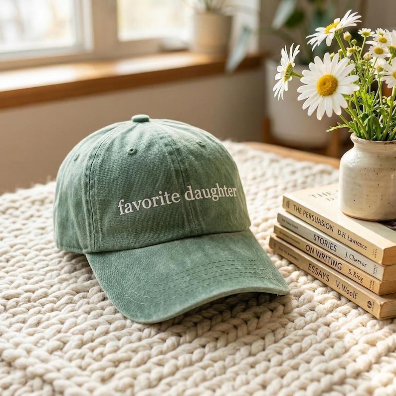 May include: A sage green baseball cap with the text "favorite daughter" embroidered in white. The hat is sitting on a cream-colored, textured surface. A stack of books and a vase of flowers are in the background.