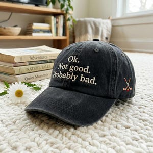 May include: Black baseball cap with the text "Ok. Not good. Probably bad." embroidered in white. The cap also features crossed hockey sticks on the side. The hat is sitting on a white, textured surface.
