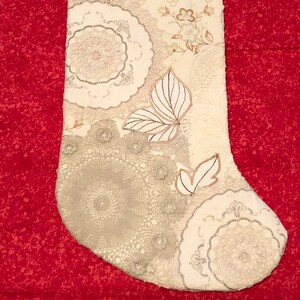 Op de afbeelding: Crèmekleurige kerstsok met verschillende kant- en bloempatronen. De sok heeft een vlinderapplicatie en een ophanglus. Het ontwerp bevat beige, crème en lichtbruine tinten.