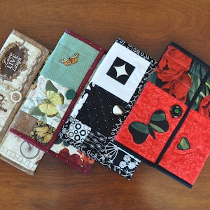 May include: Four fabric wallets with different patterns. The first wallet has a brown background with a floral pattern and the text "Love Live Laugh". The second wallet has a green background with a butterfly and floral pattern. The third wallet has a black and white background with a star pattern. The fourth wallet has a red background with a floral pattern and a clover.