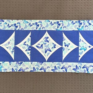 May include: A blue and white table runner with a diamond pattern and a butterfly print. The runner is made of fabric and has a quilted texture.
