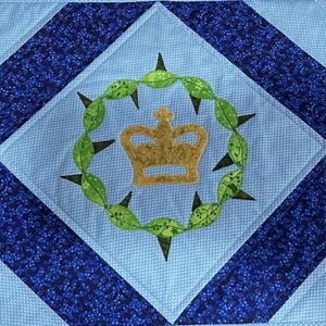 May include: A quilted square with a light blue center featuring a gold crown inside a green leafy wreath with black thorns. The center is surrounded by a white dotted light blue square, and a dark blue patterned border.