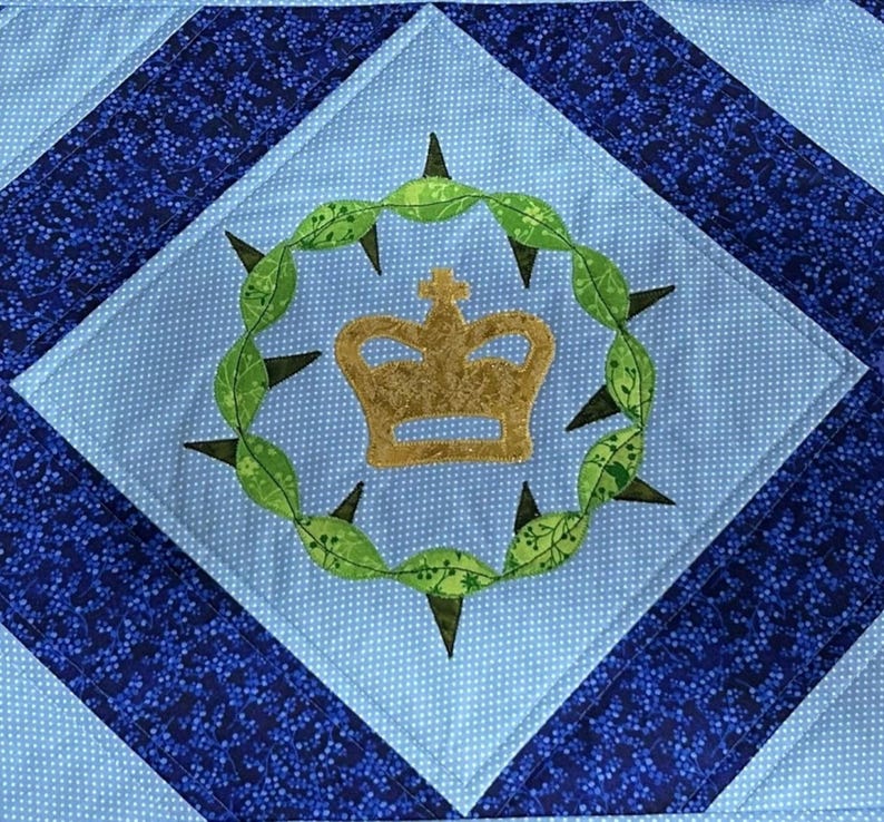 May include: A quilted square with a light blue center featuring a gold crown inside a green leafy wreath with black thorns. The center is surrounded by a white dotted light blue square, and a dark blue patterned border.