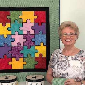 May include: A woman smiles in front of a colorful quilt made of puzzle pieces. The quilt is green, red, yellow, blue, purple, and pink. Two round boxes with black and white designs are in the foreground.