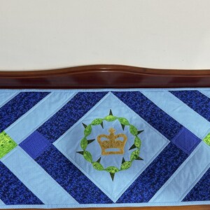 May include: A blue and navy quilted table runner with a central embroidered crown design. The runner features a diamond pattern with green and blue accents. The crown is surrounded by a green wreath.