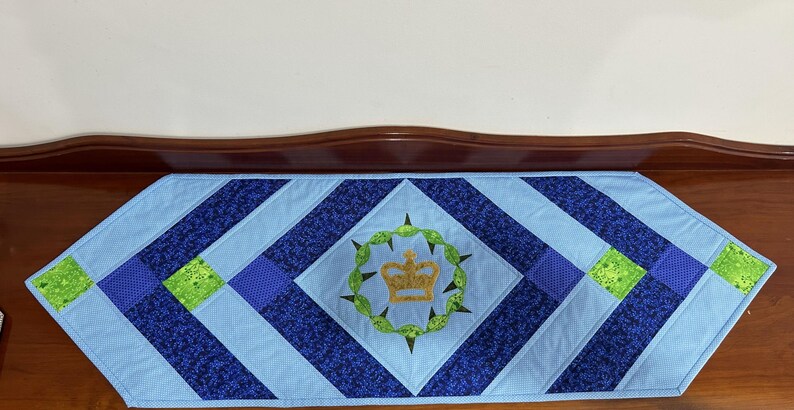 May include: A blue and navy quilted table runner with a central embroidered crown design. The runner features a diamond pattern with green and blue accents. The crown is surrounded by a green wreath.