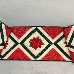Puede incluir: Un camino de mesa acolchado y dos manteles individuales a juego con un patrón geométrico de diamantes en rojo, crema y verde oscuro. Cada sección está decorada con un estampado central de flor de pascua roja y copos de nieve verdes.