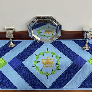 May include: A blue and navy quilted table runner with a central emblem of a gold crown within a green wreath. Silver decorative pieces are arranged on the table runner, including a mirrored tray and two small chalices.