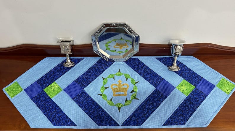 May include: A blue and navy quilted table runner with a central emblem of a gold crown within a green wreath. Silver decorative pieces are arranged on the table runner, including a mirrored tray and two small chalices.