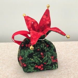 Puede incluir: Una bolsa de regalo de tela roja y verde con un patrón de acebo y una parte superior roja en forma de estrella con cascabeles dorados.