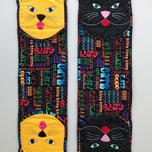 May include: Two fabric cat-shaped pillows with black and yellow cat faces. The pillows are covered in a black fabric with colorful text that says "CATS" and "KITTY" repeated many times.