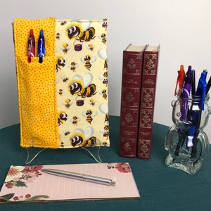 May include: A yellow and white fabric notebook cover with a bee pattern and a yellow pocket for pens. The cover is on a clear stand. There are two books, a notepad, and a pen holder with pens on a teal table.