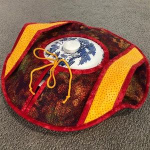 Puede incluir: Un posavasos redondo acolchado con ribete rojo y amarillo. El posavasos está decorado con un estampado floral marrón y verde. Una tapa de cerámica blanca con un diseño floral azul y blanco está colocada encima del posavasos.