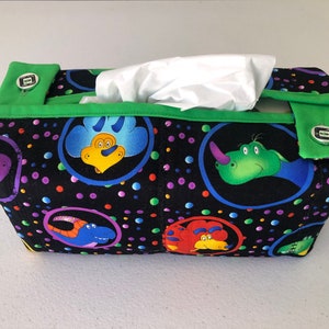 May include: A black tissue box cover with a green trim and a colorful dinosaur print. The cover has a polka dot pattern and features different dinosaurs in circles.