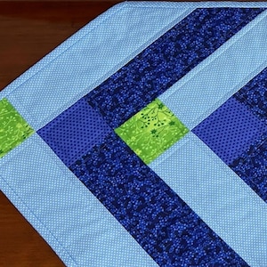 May include: A quilted table runner with a geometric design. The runner features light blue, dark blue, and green fabric squares and stripes. The light blue fabric has a polka dot pattern. The dark blue fabric has a floral pattern.