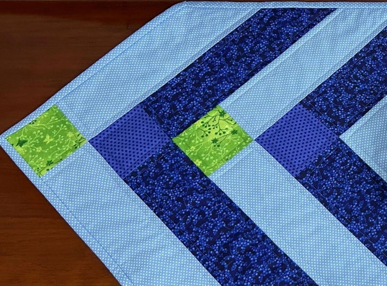 May include: A quilted table runner with a geometric design. The runner features light blue, dark blue, and green fabric squares and stripes. The light blue fabric has a polka dot pattern. The dark blue fabric has a floral pattern.