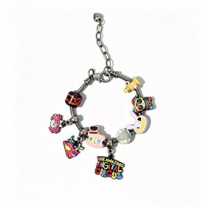 Puede incluir: Una pulsera de dijes plateada con varios dijes de personajes coloridos y un colgante central con el texto THE AMAZING DIGITAL CIRCUS en letras multicolores. Los dijes incluyen caras estilizadas, una cuenta roja y negra y un objeto amarillo.